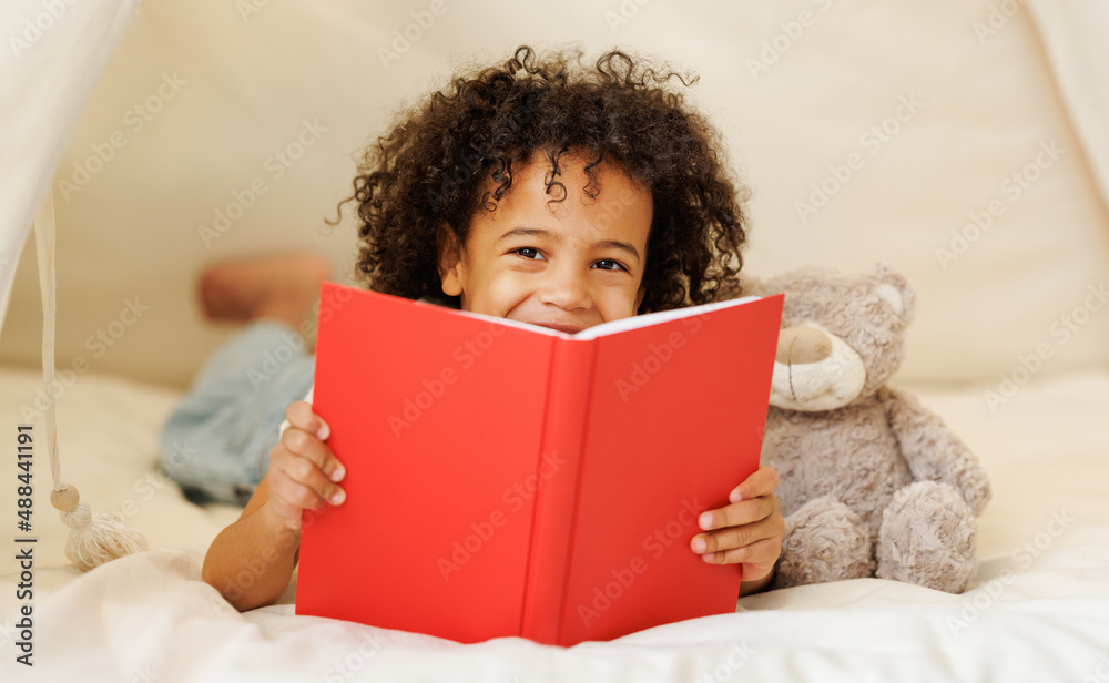 Cute little african american kid curly boy holding book lying in play tent with teddy bear toy