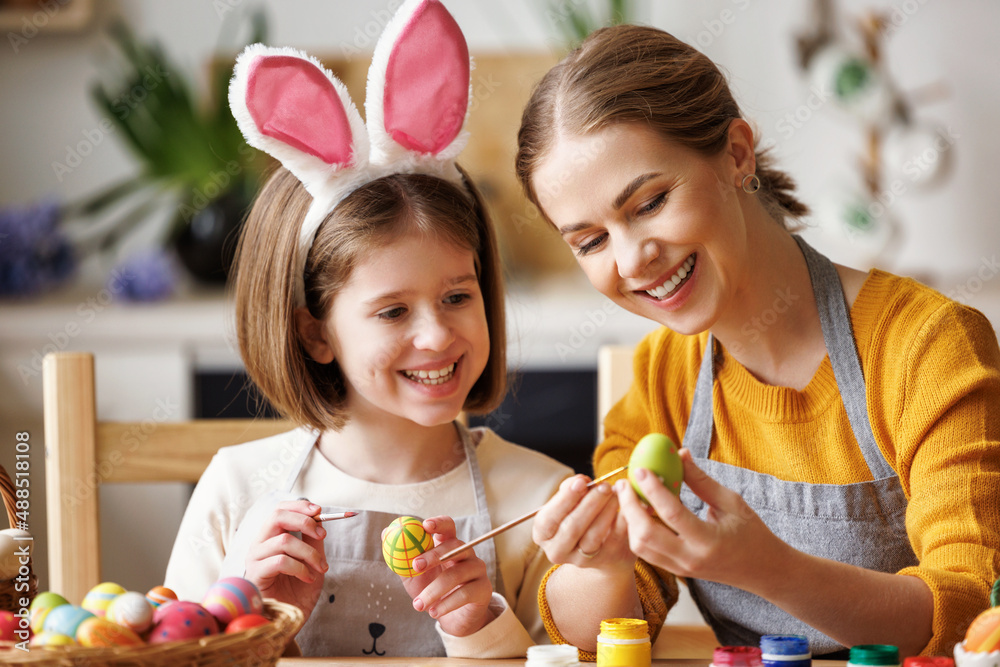 慈爱的年轻母亲坐在厨房里教快乐的小女儿装饰复活节彩蛋