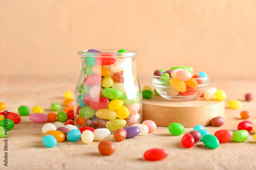 Jar and bowl with different jelly beans on beige background, closeup
