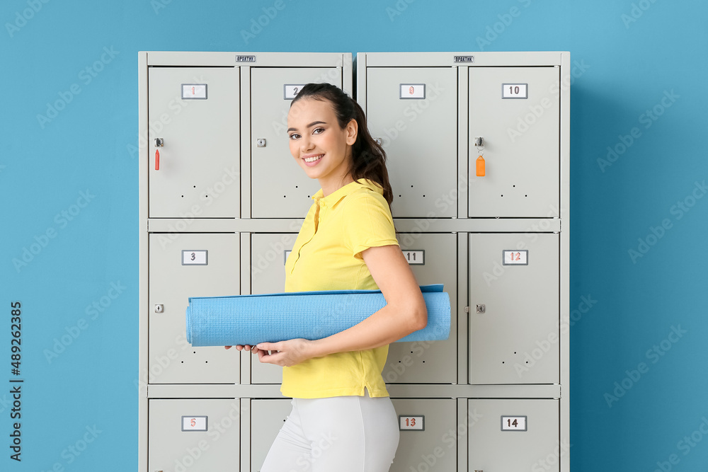 蓝色背景储物柜附近有一个带瑜伽垫的运动型年轻女子