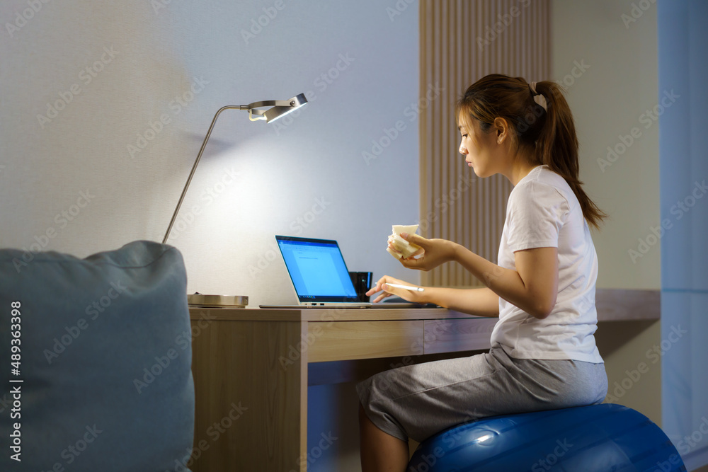 亚洲女性在瑜伽球上放松，吃三明治，并在网上与同事聊天