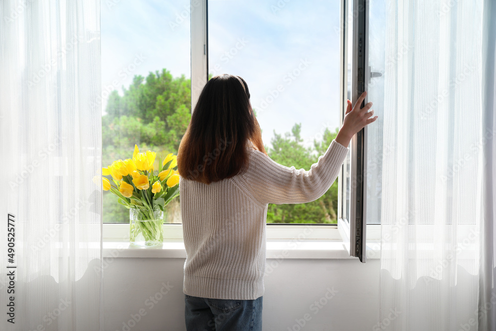 阳光明媚的日子里，漂亮的年轻亚洲女人在家里开窗，背影