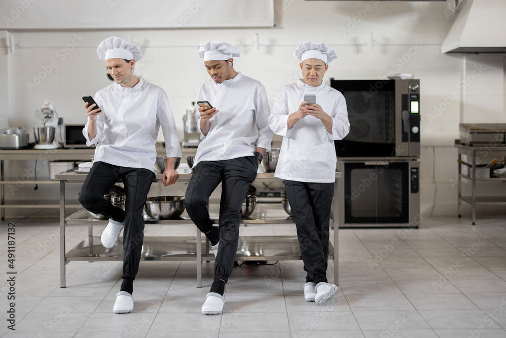 三名多种族厨师在餐厅厨房休息时拿着智能手机站着。As