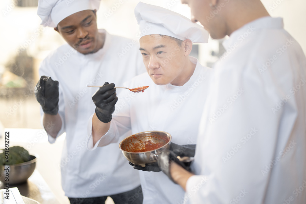 三位不同种族的厨师在厨房做饭时用勺子品尝酱汁。