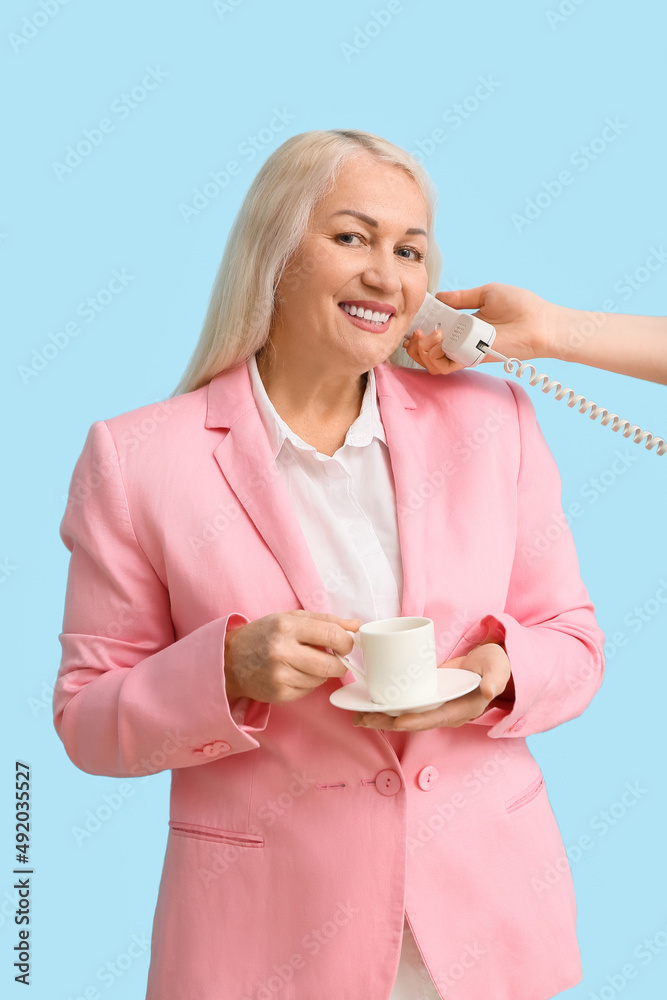 Mature secretary with cup of coffee talking by phone on blue background