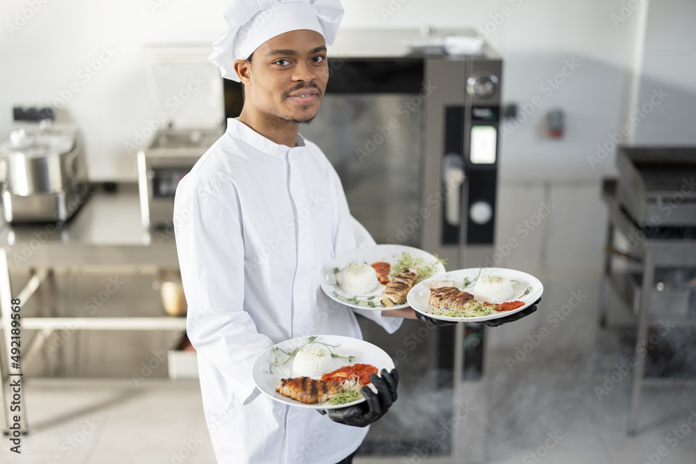 身着制服、面带微笑的年轻英俊厨师站在厨房里准备好饭菜的肖像。开心啦