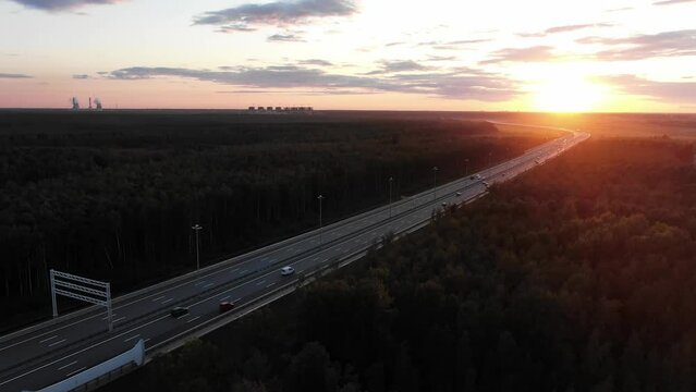 Long highway with driving cars running among woods with river stretches towards setting sun in summer evening aerial panorama
