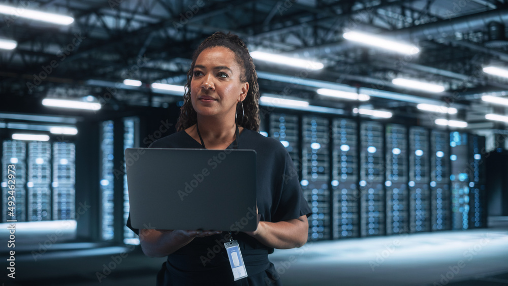 站在数据中心，成功的非裔美国女性IT专家使用笔记本电脑的画像。