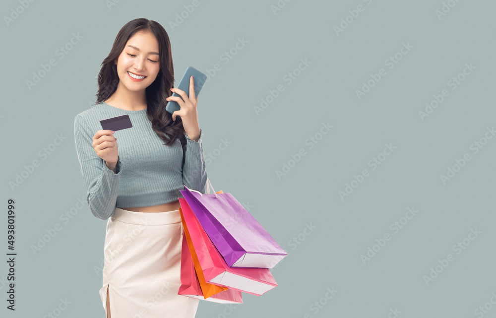 年轻的亚洲女士用手机拿着信用卡，背着被隔离在粉彩gre上的购物袋
