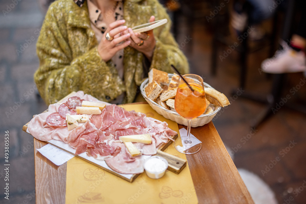 坐在餐厅时，女人拿着意大利肉类开胃菜和雪碧Aperol饮料拍照