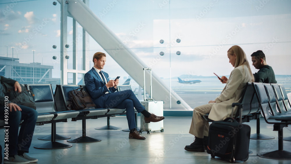 繁忙的机场航站楼：英俊的商人在等待航班时使用智能手机。人们坐着
