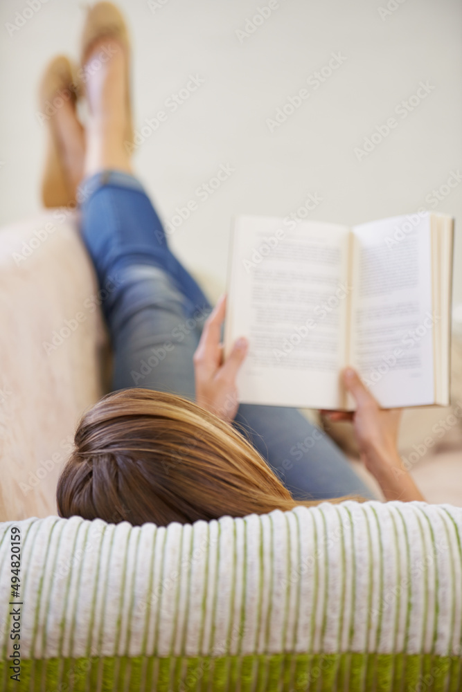 读一本好书放松一下。一个女人躺在家里的沙发上看书的照片。