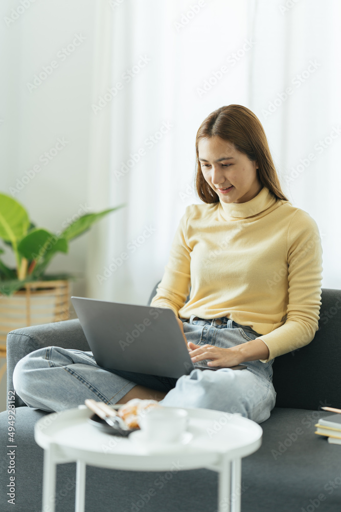 Smiling beautiful young asian ethnicity woman sitting on sofa with computer on lap, thinking of dail