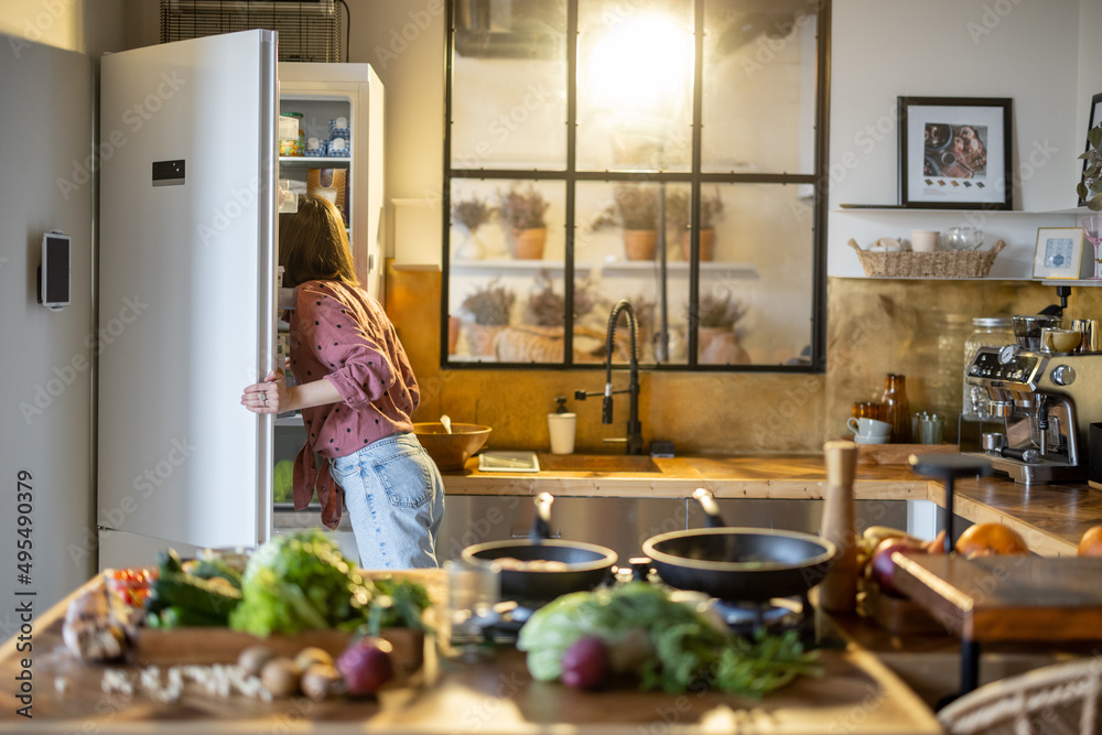 Woman looks into a fridge while cooking in the kitchen at home, healthy food on table top in front. 