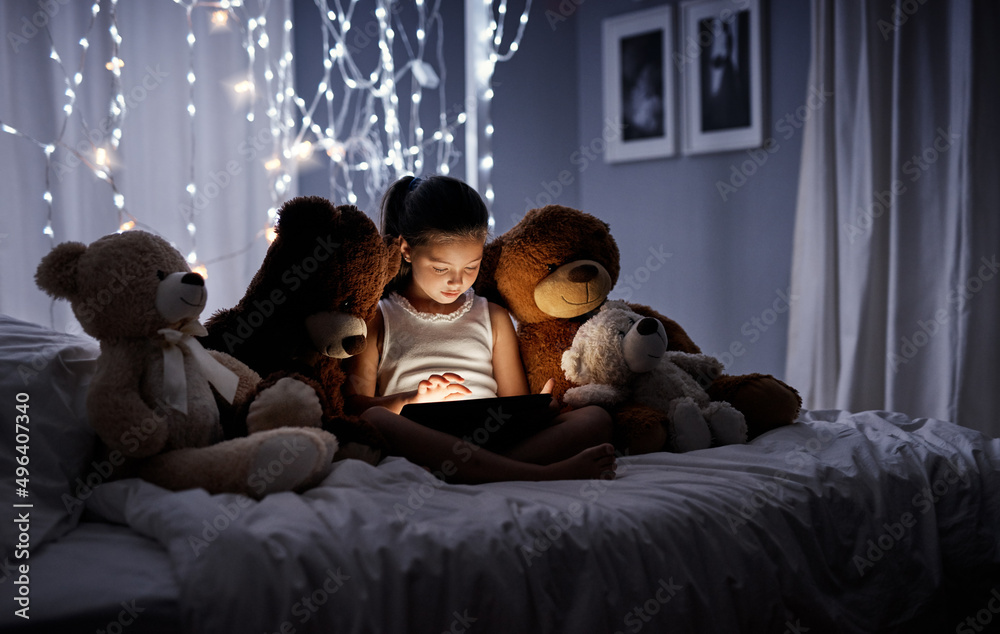 向技术介绍她的泰迪熊。一个可爱的小女孩在床上使用数字平板电脑的照片
