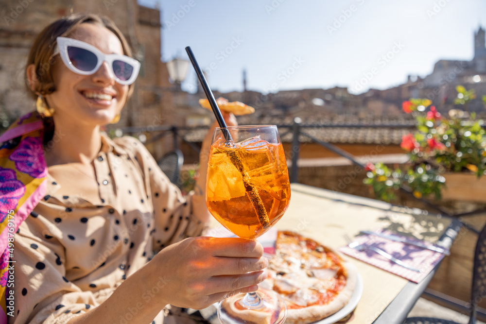 Young woman cheering at camera with a glass of wine, while eating pizza at outdoor restaurant in Sie