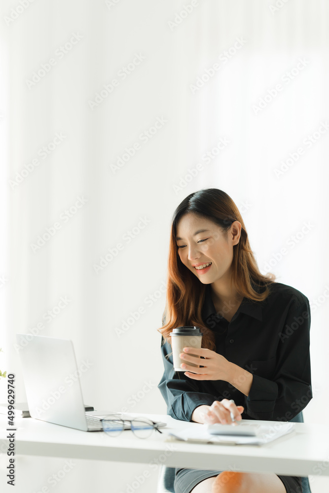 漂亮的年轻亚洲商务女性在办公室工作时喝咖啡并使用笔记本电脑