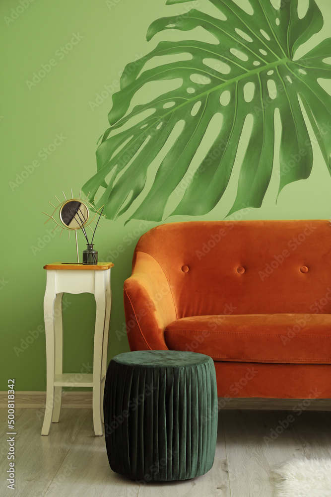 带桌子和脚凳的时尚沙发，靠近绿色墙壁，画着热带树叶