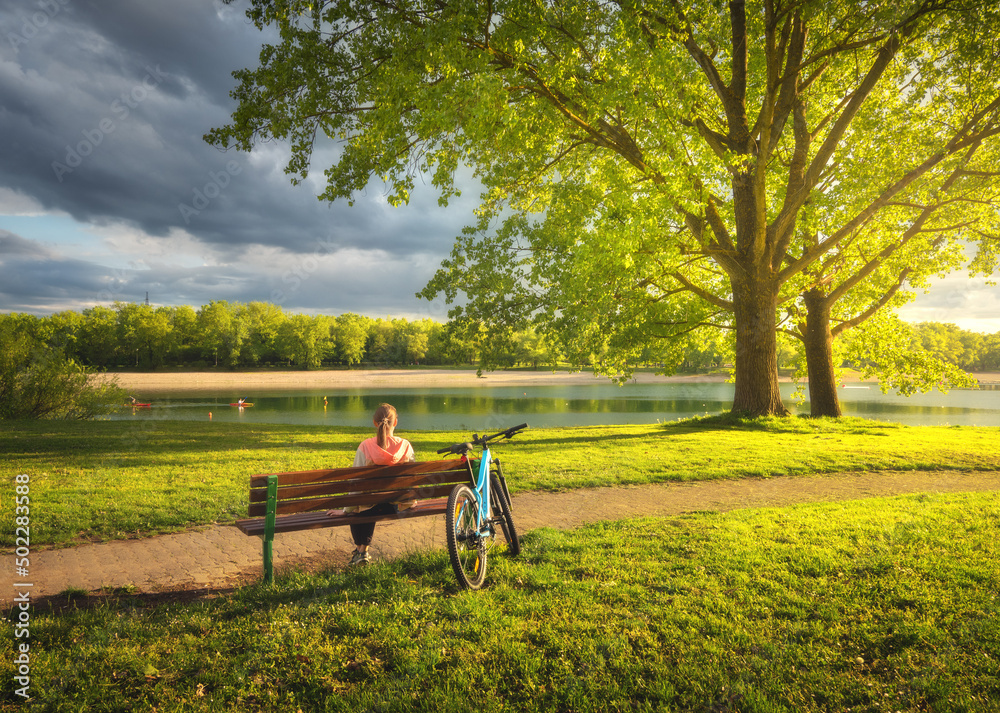 Woman sitting on bench and mountain bike, green trees and lake at sunset in spring. Colorful landsca