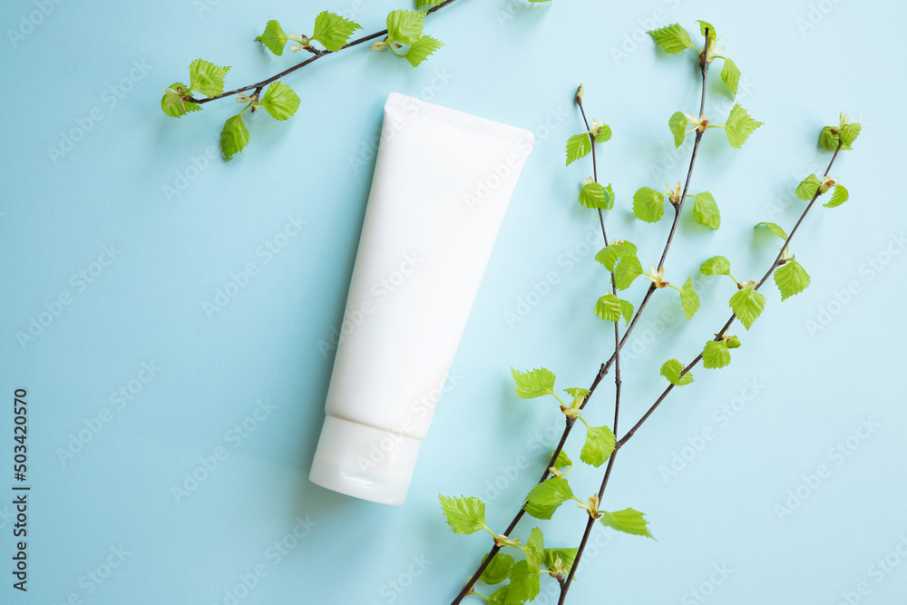 White Cream tube and birch branches with young small leaves on blue background. Cosmetic skincare pr