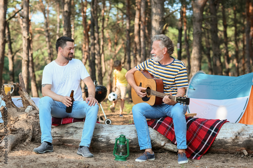 一名老人在森林里弹吉他，他的儿子拿着一瓶啤酒