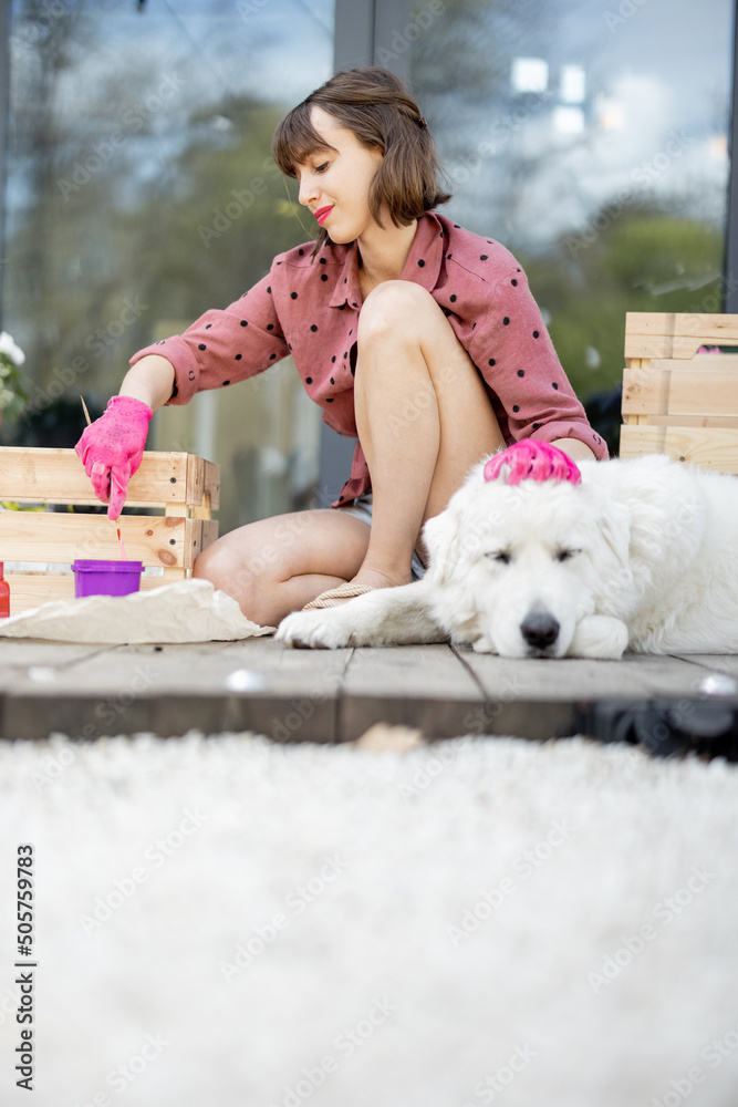 Young woman painting wooden box in pink color, doing housework while sitting with her dog on terrace