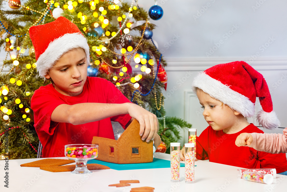 两个孩子戴着圣诞老人帽一起装饰姜饼屋