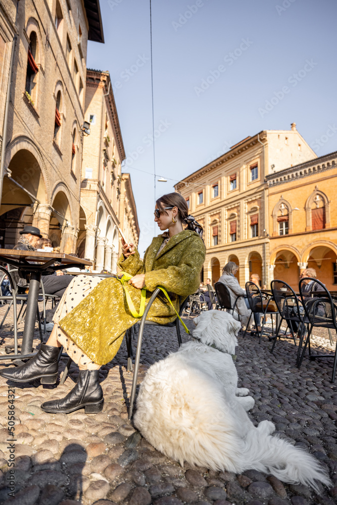 时尚的女人和她的白狗坐在博洛尼亚老城区的户外咖啡馆里。意大利的我