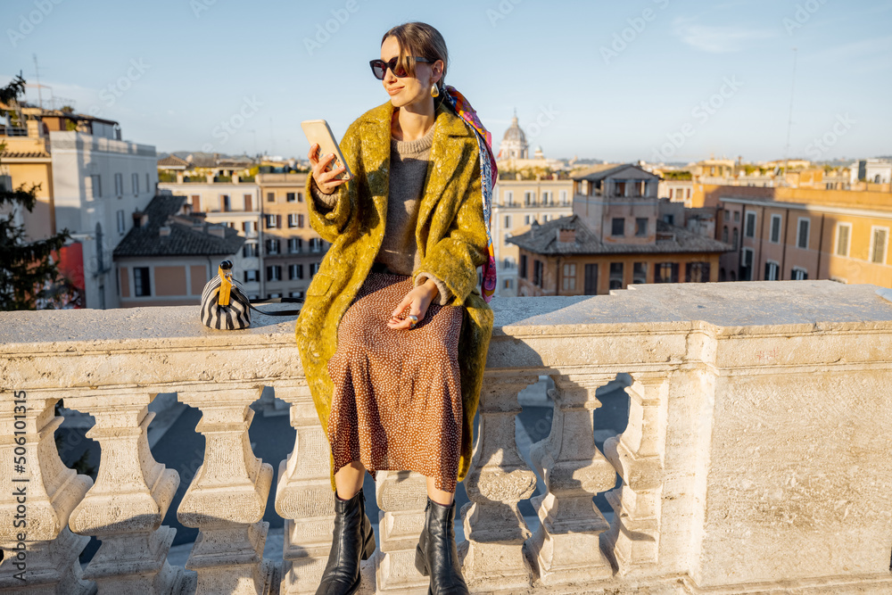 一位女士坐在屋顶上，享受着罗马美丽的早晨城市景观，在手机上拍照