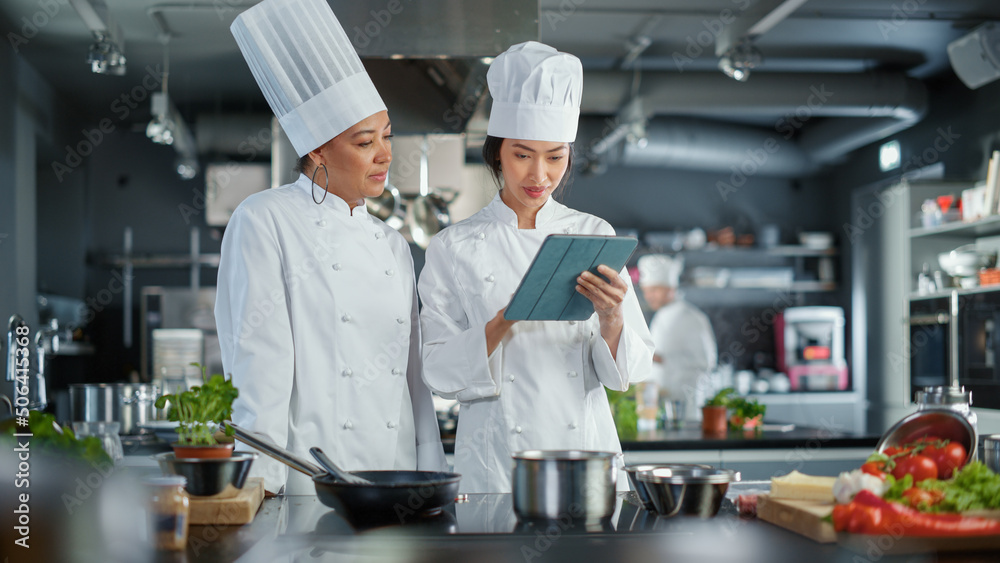 Restaurant Kitchen: Team of Asian and Black Female Chefs use Tablet Computer while Cooking Delicious
