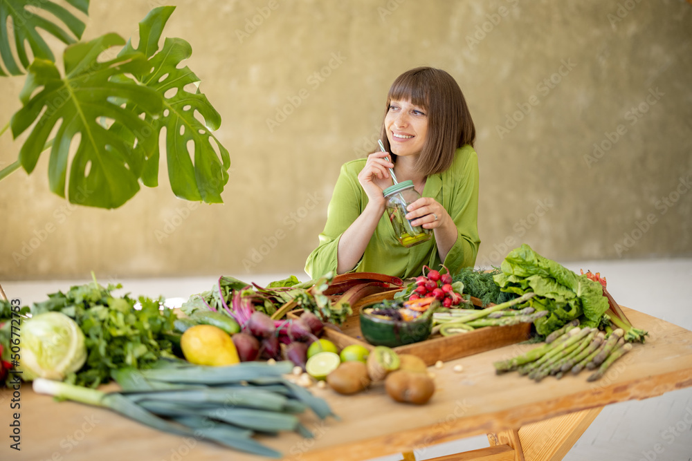 Young woman drinks from a bottle while sitting by the table full of fresh vegetables, fruits and gre