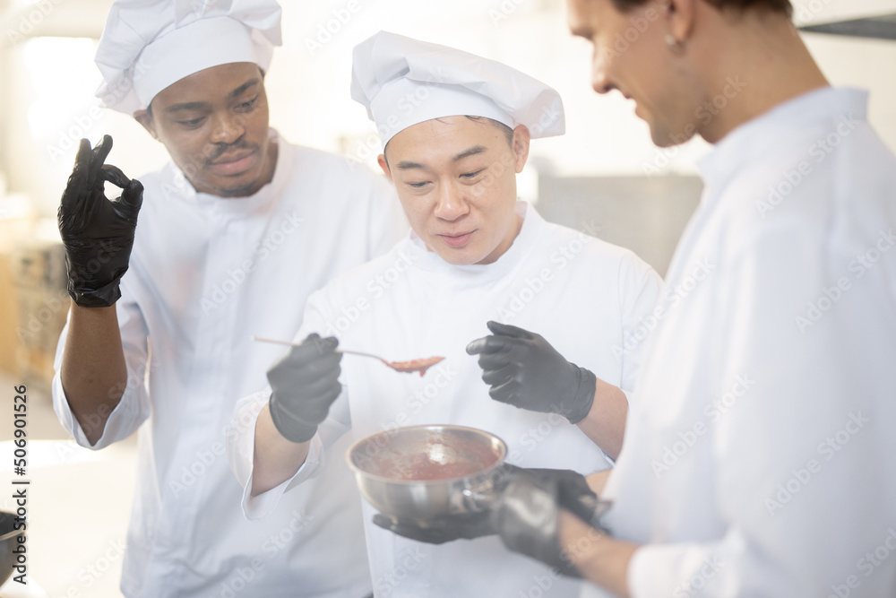 Three chef cooks with different ethnicities tasting sauce with a spoon while cooking in the kitchen.