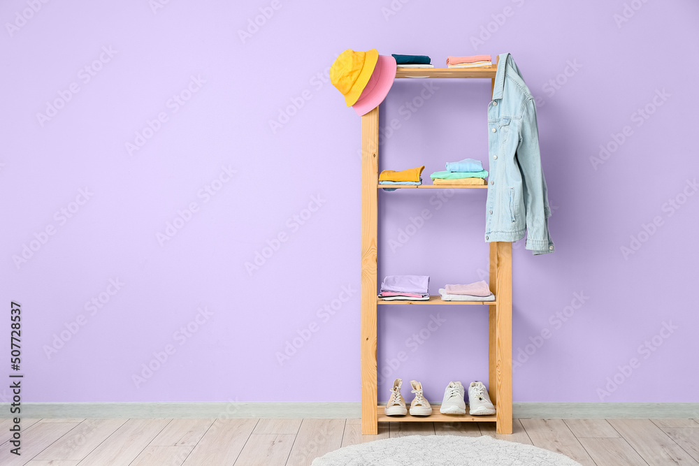 Wooden shelf unit with stylish clothes and shoes near violet wall in room interior