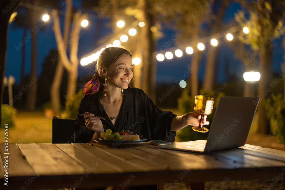 微笑的高加索女性视频通话，对着笔记本电脑敬酒。坐在桌子旁的年轻女性