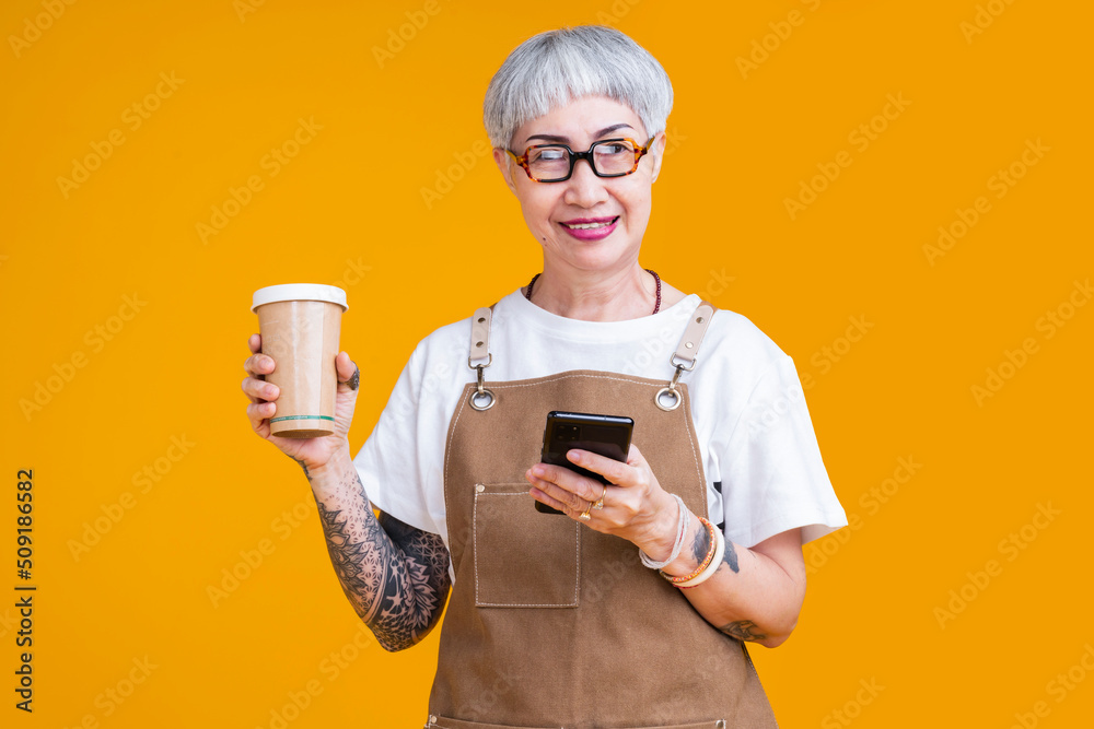 资深老年女性纹身企业主系围裙手持咖啡杯和智能手机人像微笑