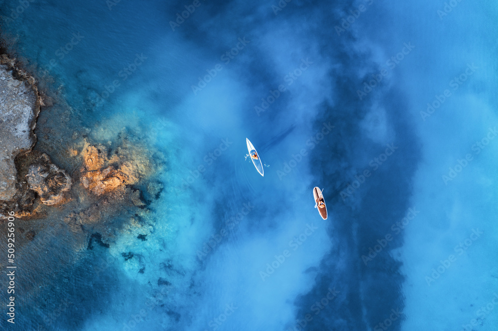 蓝色海洋、岩石海岸上漂浮的皮划艇上的人们的鸟瞰图，夏天日出时的树木。蓝色