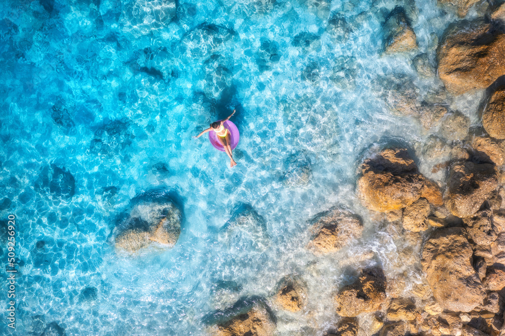 夏日日落时，一名年轻女子戴着粉色游泳圈在蓝色大海中游泳的鸟瞰图。热带