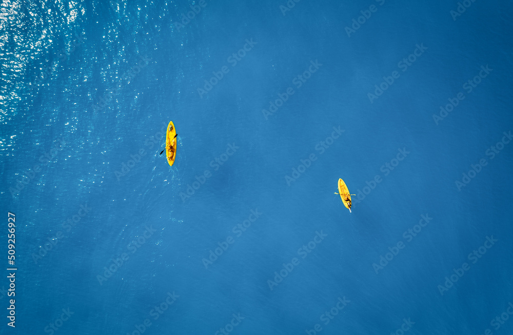 夏日日落时，蓝色大海中黄色皮划艇的鸟瞰图。男人和女人在独木舟上漂浮