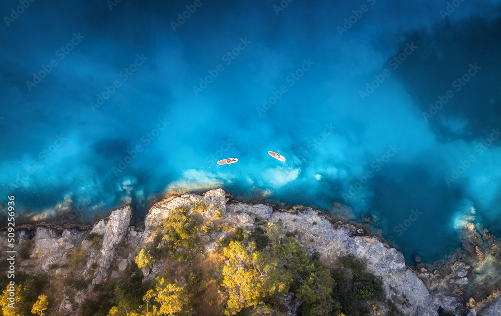 蓝色海洋、岩石海岸、夏季日落时树木上漂浮的皮划艇上的人们的鸟瞰图。蓝色l