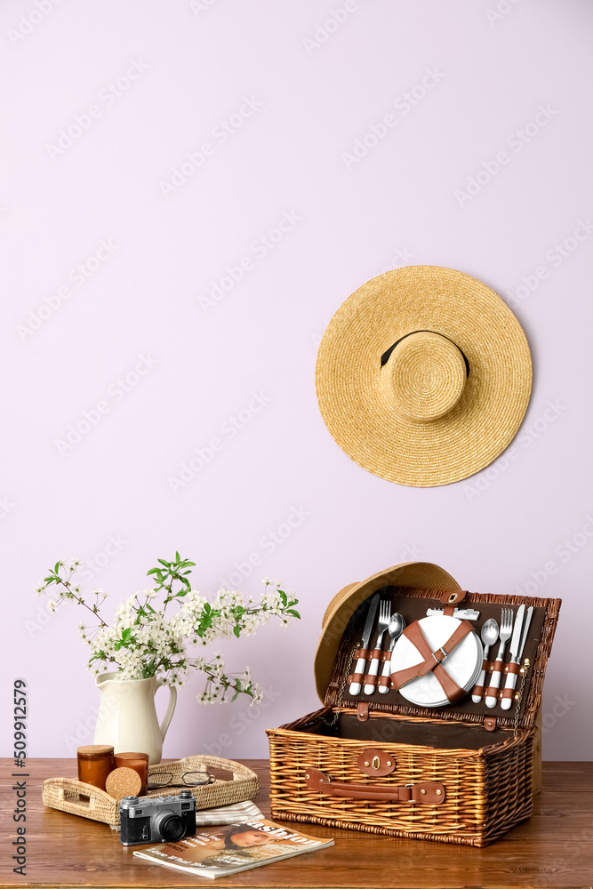 Basket with dishware, photo camera, magazine and blooming branches in vase on table near light wall