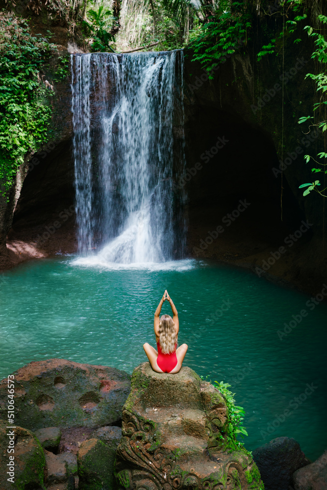 身穿比基尼的女子坐在岩石上，在热带苏瓦特瀑布的瀑布下以瑜伽姿势冥想