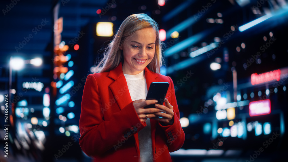 美丽的女人站在城市街道上，使用智能手机，夜晚霓虹灯闪烁。C