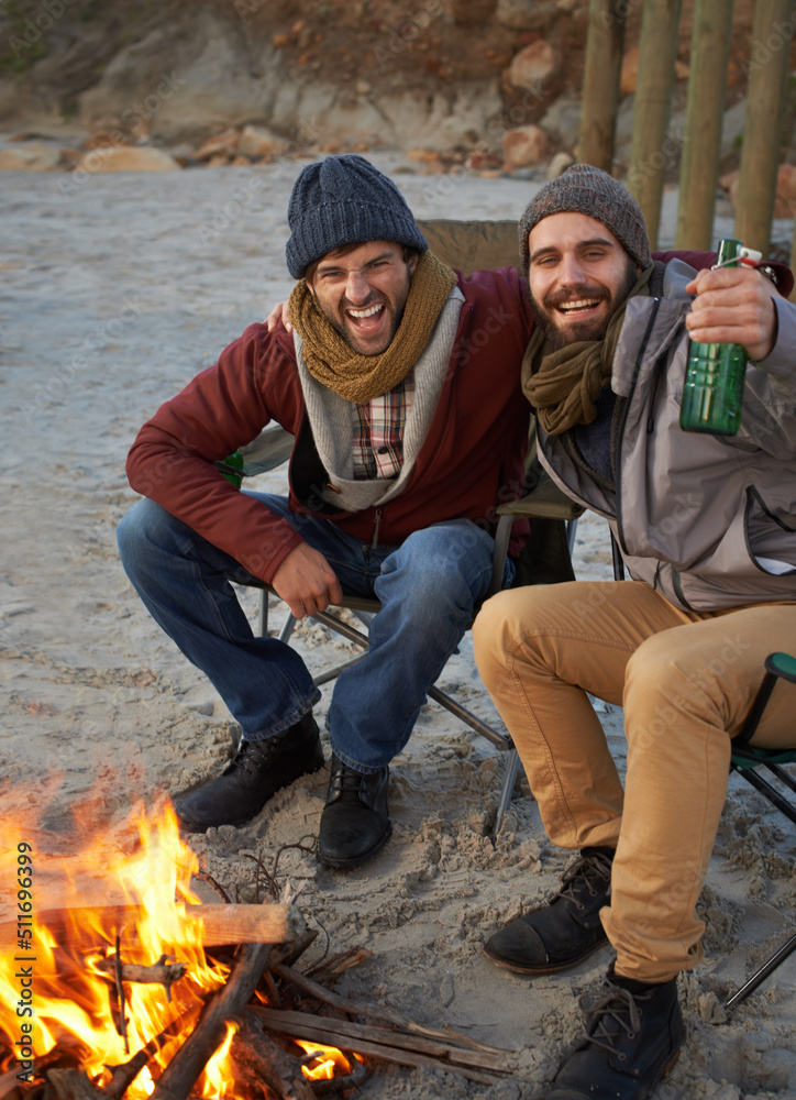 用炉火和啤酒取暖。两个年轻人坐在海滩上的炉火旁。