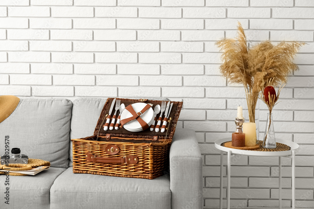Sofa with basket for picnic, photo camera, bag and table near white brick wall