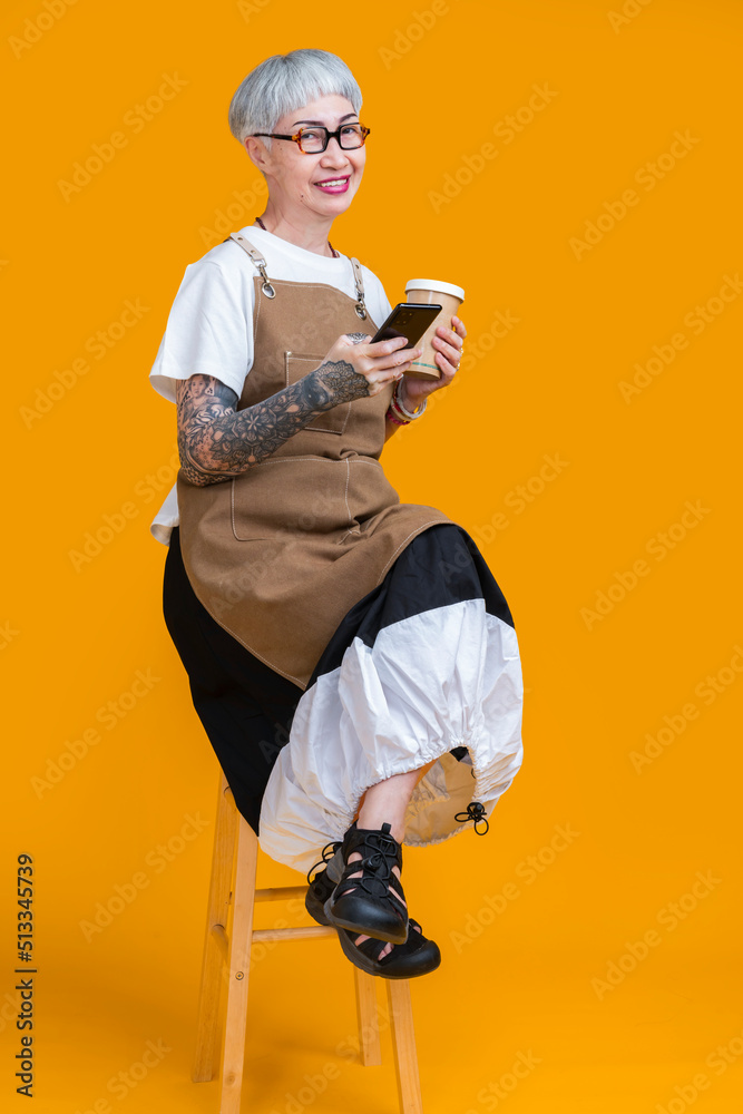 资深老年女性纹身企业主系围裙手持咖啡杯和智能手机人像微笑
