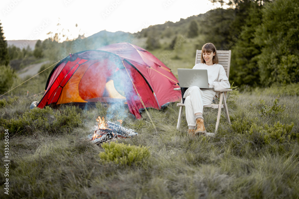 一名年轻女子一边用笔记本电脑工作，一边放松地坐在篝火旁的椅子上，带着帐篷旅行