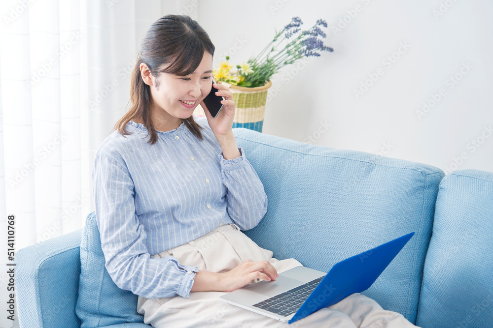 リビングでノートパソコンを見てスマホで電話する女性