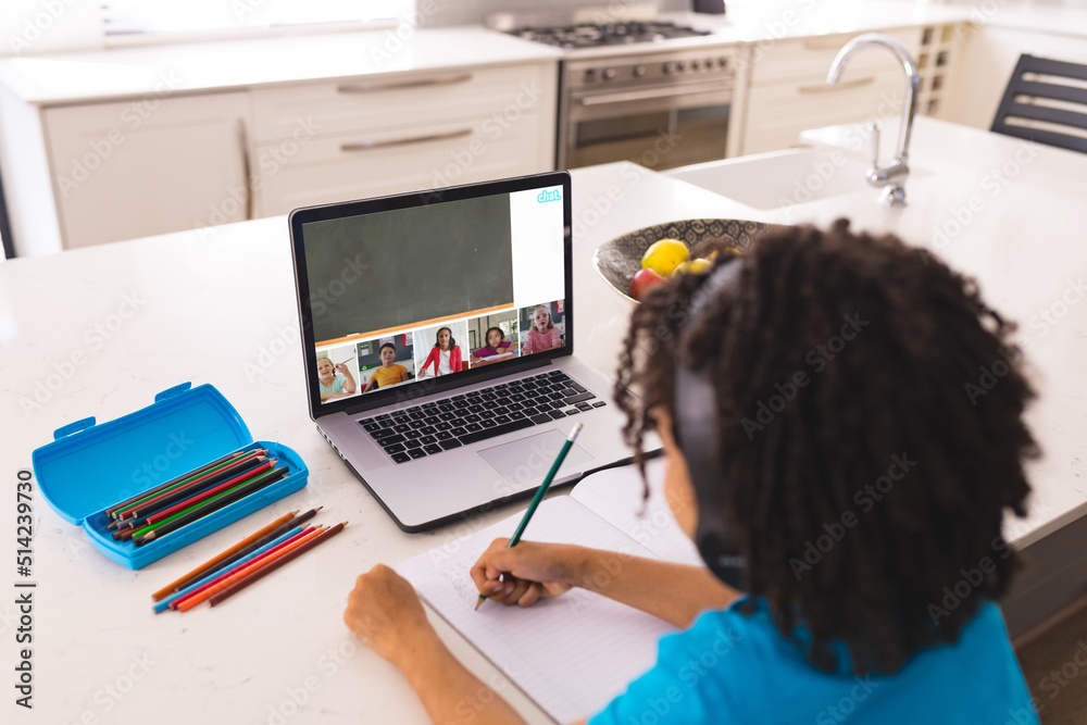 一个留着卷发的非裔美国男孩在家里桌子上的笔记本电脑上在线学习时写作