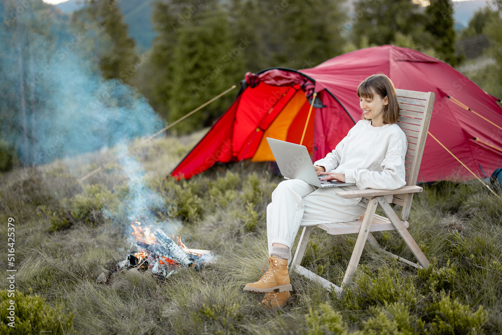 一名年轻女子在篝火旁的椅子上放松地坐着，带着帐篷旅行，一边用笔记本电脑工作