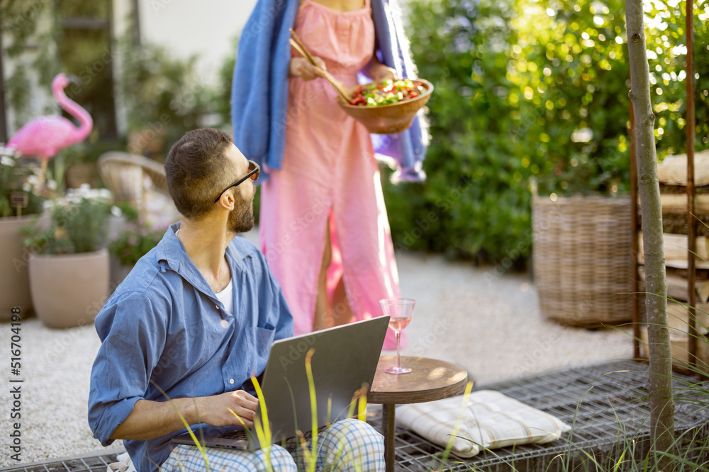 男人坐在后院用笔记本电脑工作。女人带着健康沙拉。远程工作的概念